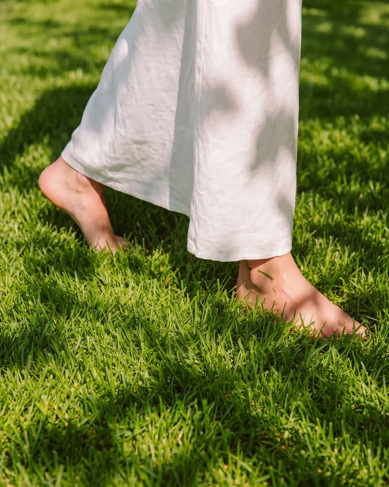 Bare feet mid-stride on thick sunlit grass, under the hem of white linen trousers.