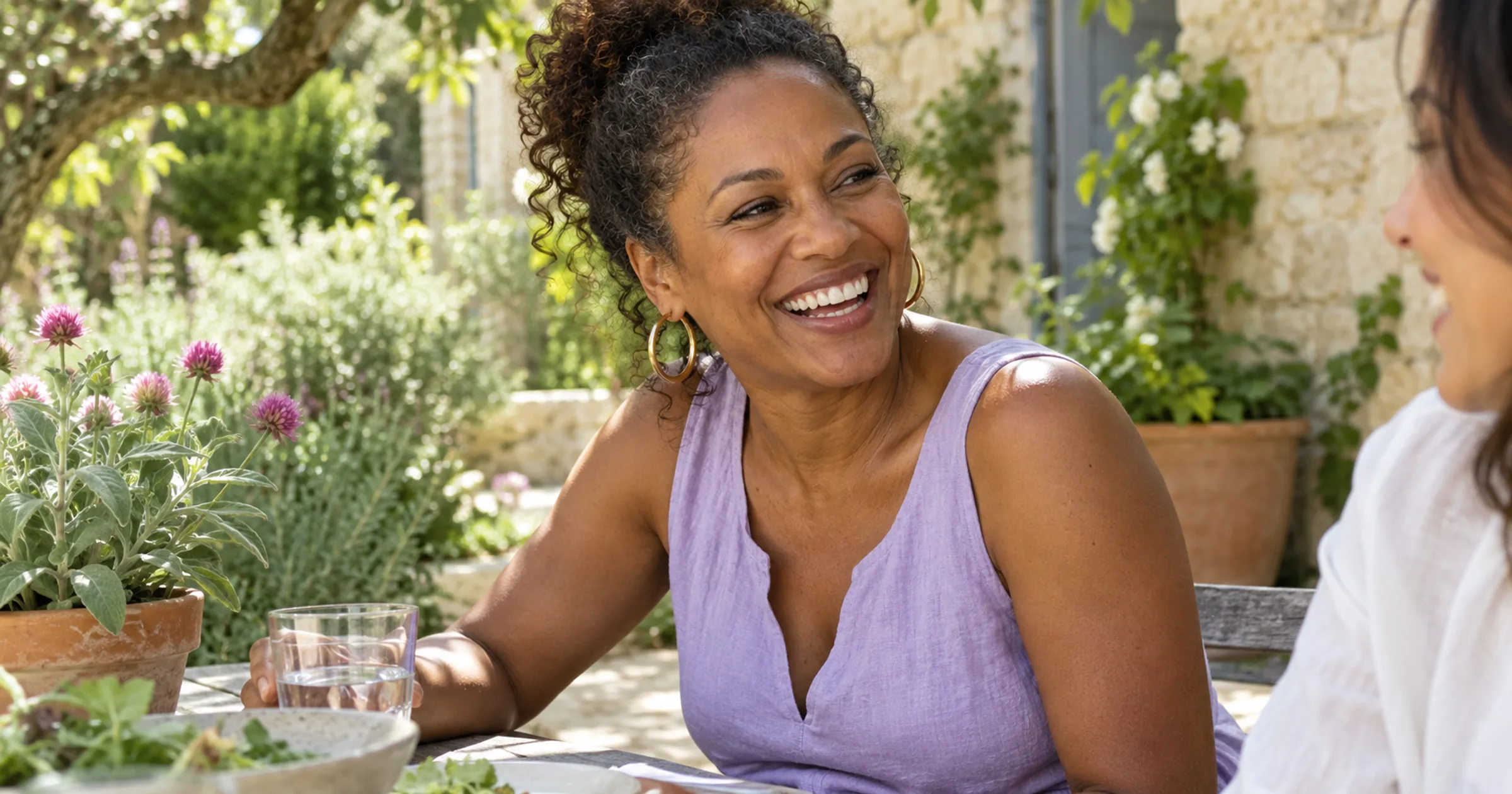 A woman in a lavender linen top laughing with a friend at a garden table beside sage and red clover flowers.